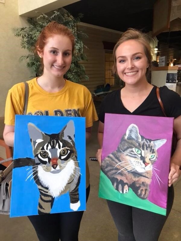 dos chicas cada una asegurando un lienzo con una pintura de gato.